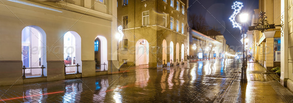 Panorama of night street in Klaipeda, Lithuania. Long narrow street paved with stone with illuminated buildings along it. Wet road glittering after rain with reflection from light of windows.