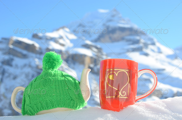 Tea pot in the knitted cap and red cup with a heart in the snow