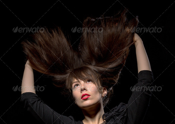 Young woman with her long straight brunette hair flying in the air and her arms raised above her head isolated on black