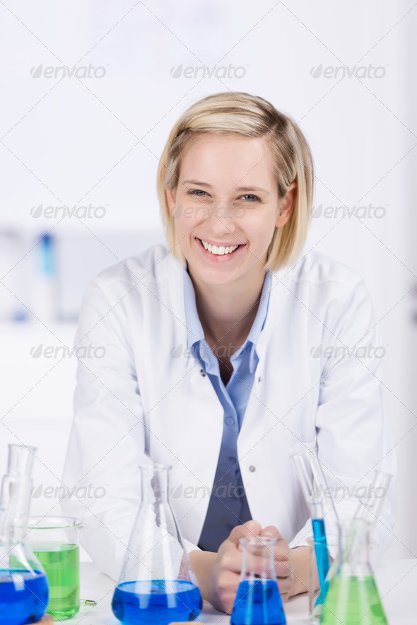 Happy laboratory technician or technologist standing behind a counter filled with chemical solutions and glassware looking at the camera with a beautiful smile on her face