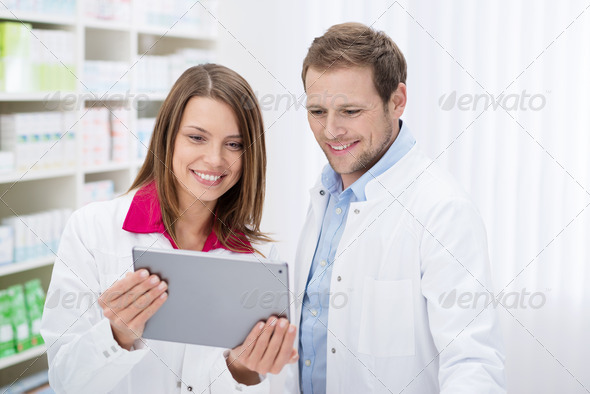 Two smiling young pharmacists, one male and one female, stand side by side in the pharmacy checking information on a tablet computer