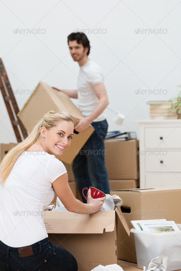 Pretty blond woman packing up the house as she and her husband get ready to move to a new home turning to smile at the camera as she kneels on the floor with a cardboard box