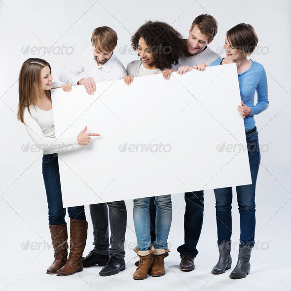 Group of young friends in trendy winter fashion standing looking at a blank sign that they are holding as the young woman on the end points to it with her finger drawing their attention to copyspace